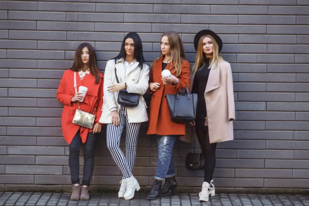Five women standing casually against a gray brick wall, dressed in stylish coats exuding a trendy vibe.