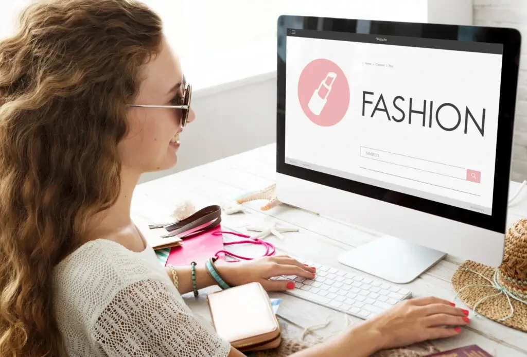 A person with long hair sits at a desk, browsing a fashion website on a computer, surrounded by accessories.
