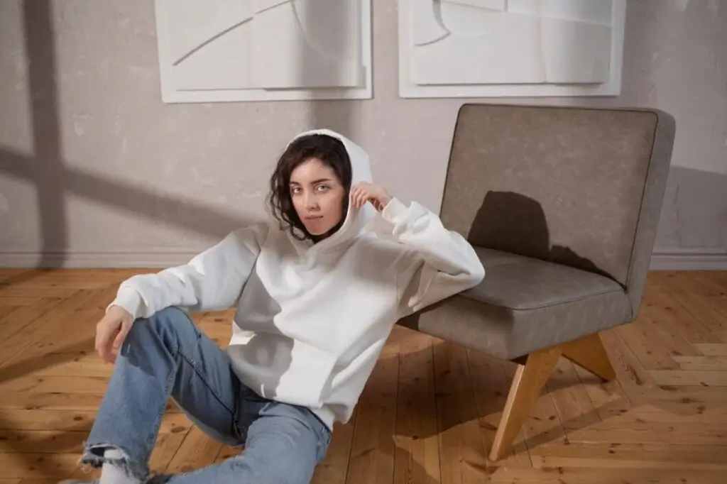 A person in a white hoodie sits casually on the floor beside a modern gray chair, with minimalist art on the wall.