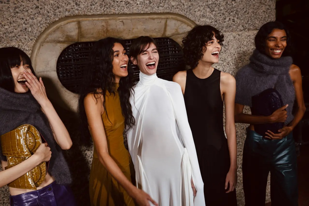Four joyful women in chic outfits laughing together near a textured stone wall.