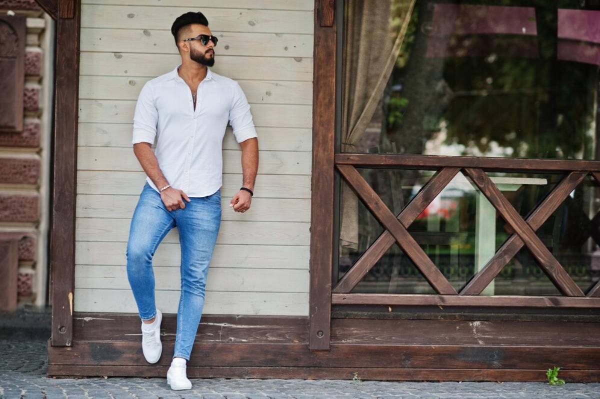 Man in white shirt and blue jeans leaning against a wooden door, wearing sunglasses.